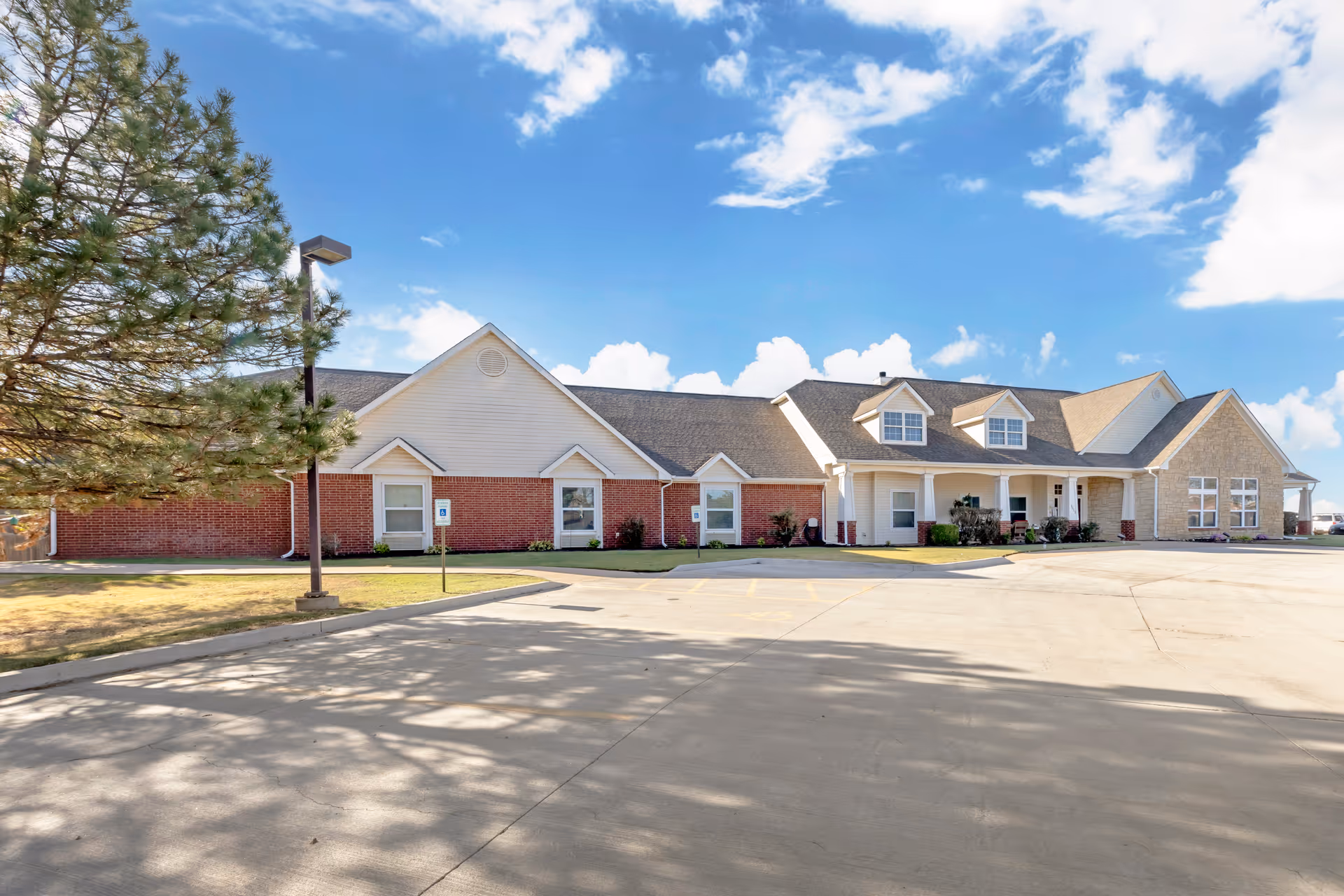 Exterior view of a single-story senior living facility building with a combination of brick, siding, and stone facade under a blue sky with scattered clouds. There is a paved driveway and parking area in front, with a tree and a street lamp on the left side.