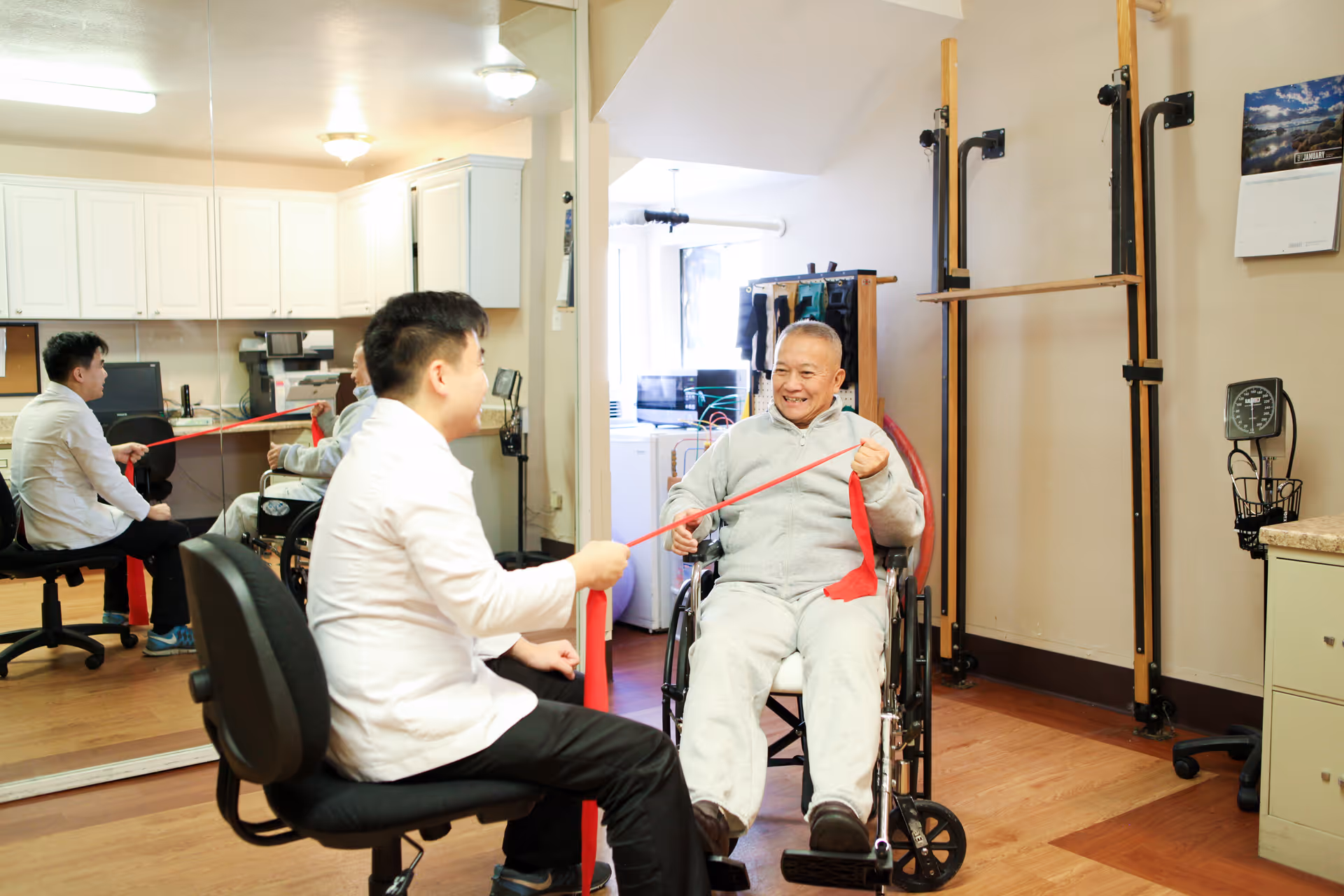 A healthcare professional in a white coat assists an elderly man in a wheelchair with a red resistance band exercise in a room with wooden floors, medical equipment, and a large mirror reflecting their image.
