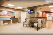Interior view of a senior living facility reception area with a front desk on the left displaying a sign that reads 'Rochester Manor + Villa.' Two upholstered chairs and a small round table are positioned in front of a stone accent wall with a mounted flat-screen TV. The area is well-lit with ceiling lights and has a clean, welcoming atmosphere.