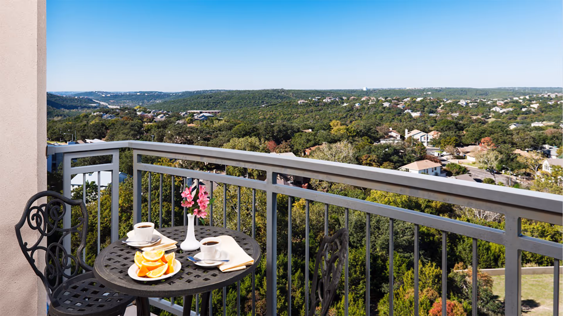 Balcony with a small table set for coffee and fruit and two chairs overlooking a tree-covered suburban landscape.