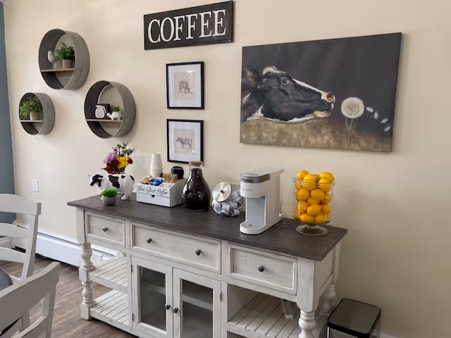 A coffee station in a senior living facility with a white wooden sideboard holding a coffee maker, a glass container filled with lemons, a dark bottle, and a small white box labeled 'The Best Coffee'. Above the sideboard are three round wall shelves with small plants and decor, two framed pictures, a large painting of a cow looking at a dandelion, and a sign that reads 'COFFEE'. A white chair is partially visible on the left side.