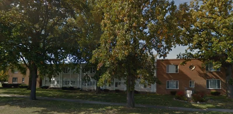 Exterior view of a two-story senior living facility building partially obscured by large trees with green foliage. The building has a combination of brick and white siding with multiple windows. A sidewalk runs in front of the building across a grassy area.