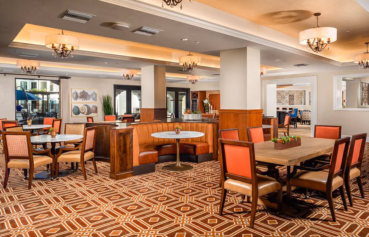 Bright, elegant dining room with several round and rectangular tables, red-upholstered chairs, a curved leather banquette, decorative chandeliers, and patterned carpeting.