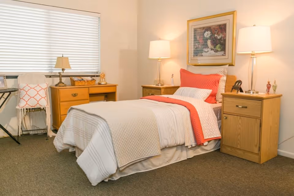 A cozy bedroom in a senior living facility featuring a single bed with white and coral bedding, two wooden nightstands with lamps, a wooden desk with a lamp and decorative items, a chair with a white throw and coral pillow, and a framed floral painting on the wall.