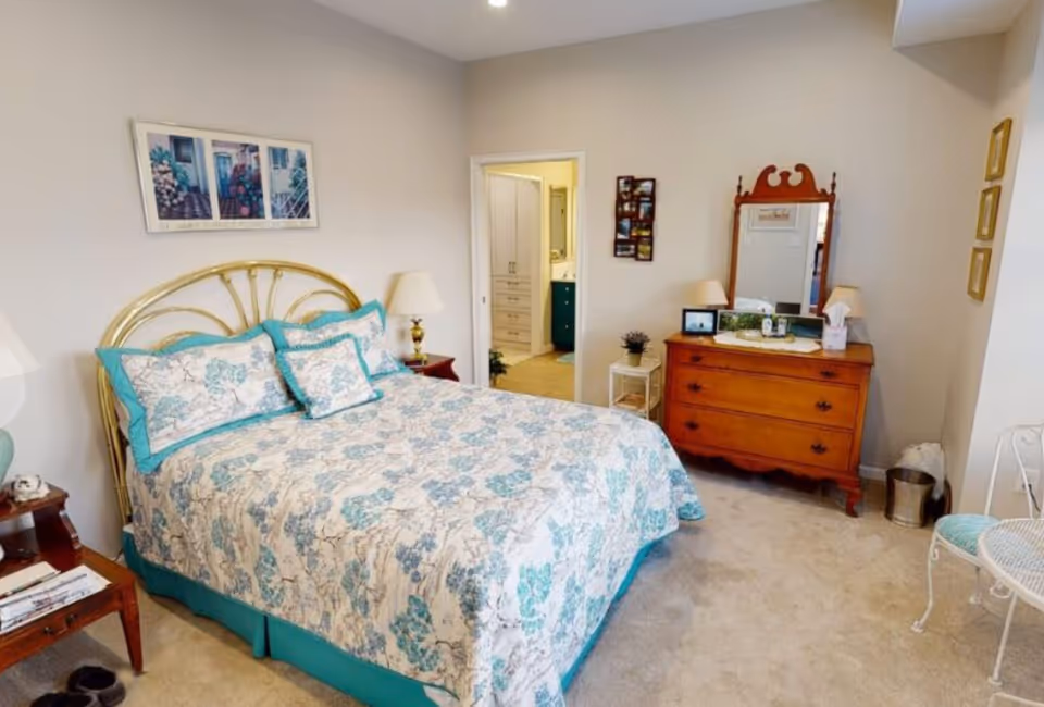 A cozy bedroom featuring a bed with a floral patterned bedspread in shades of blue and white, a brass headboard, and matching pillows. There are two bedside tables with lamps on either side of the bed. A wooden dresser with a mirror and two lamps sits against the wall, along with a small side table holding a plant. The room has beige carpeting and light-colored walls with framed artwork. An open doorway leads to a bathroom area.