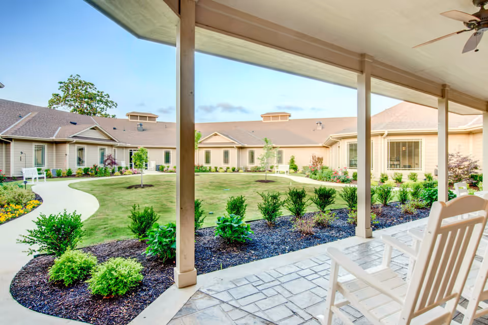 View from a covered patio with white rocking chairs overlooking a landscaped courtyard with a curved walkway, green grass, shrubs, and a beige single-story building under a clear blue sky.