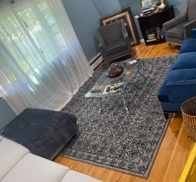 Cozy living room with a blue sofa and armchairs around a glass coffee table on a patterned rug, sheer curtains and hardwood floors.