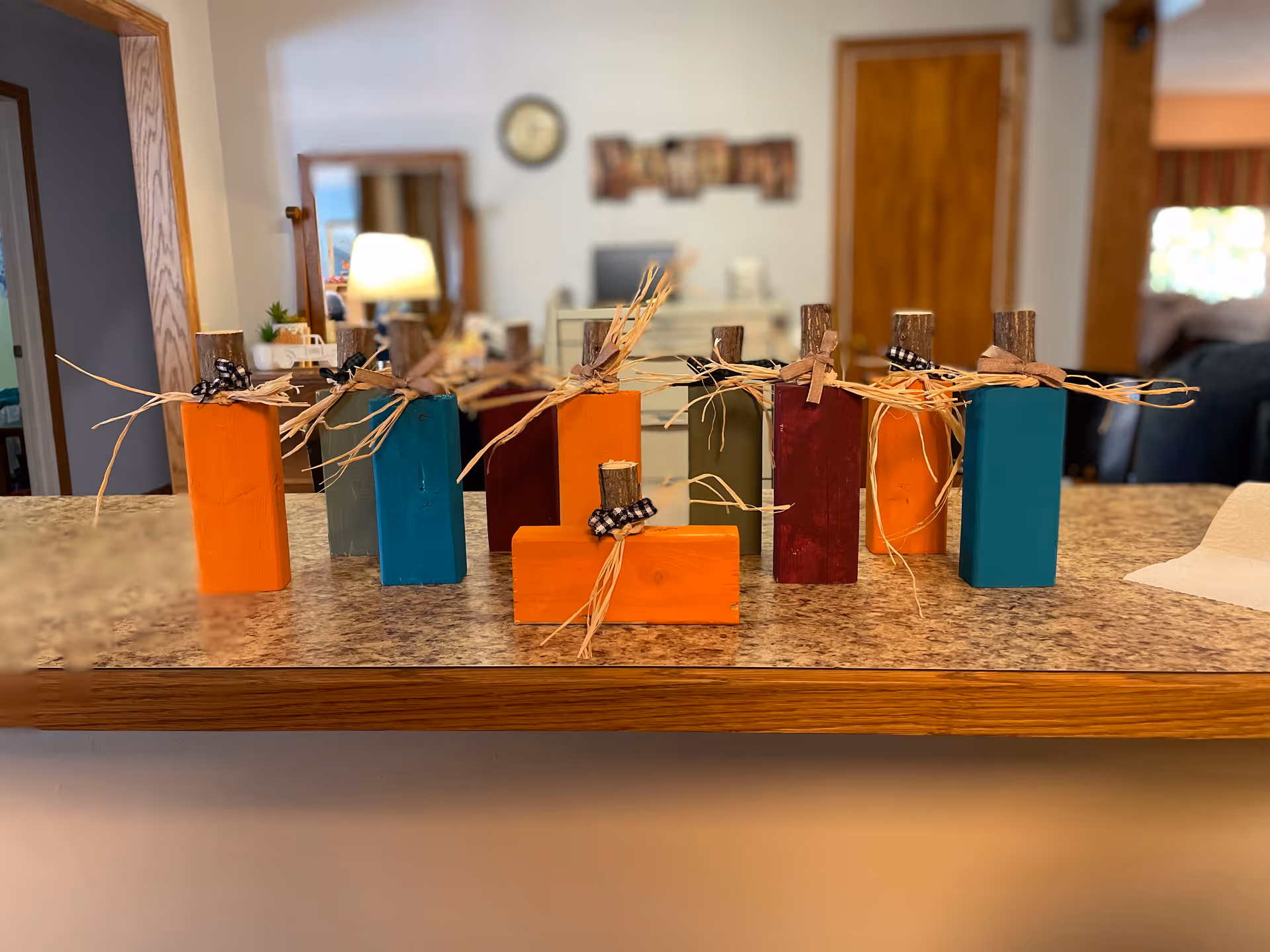 A row of colorful wooden block decorations shaped like pumpkins with small wooden stems and tied with raffia and black-and-white checkered ribbons, displayed on a kitchen counter with a blurred background of a room featuring a mirror, clock, and wooden doors.