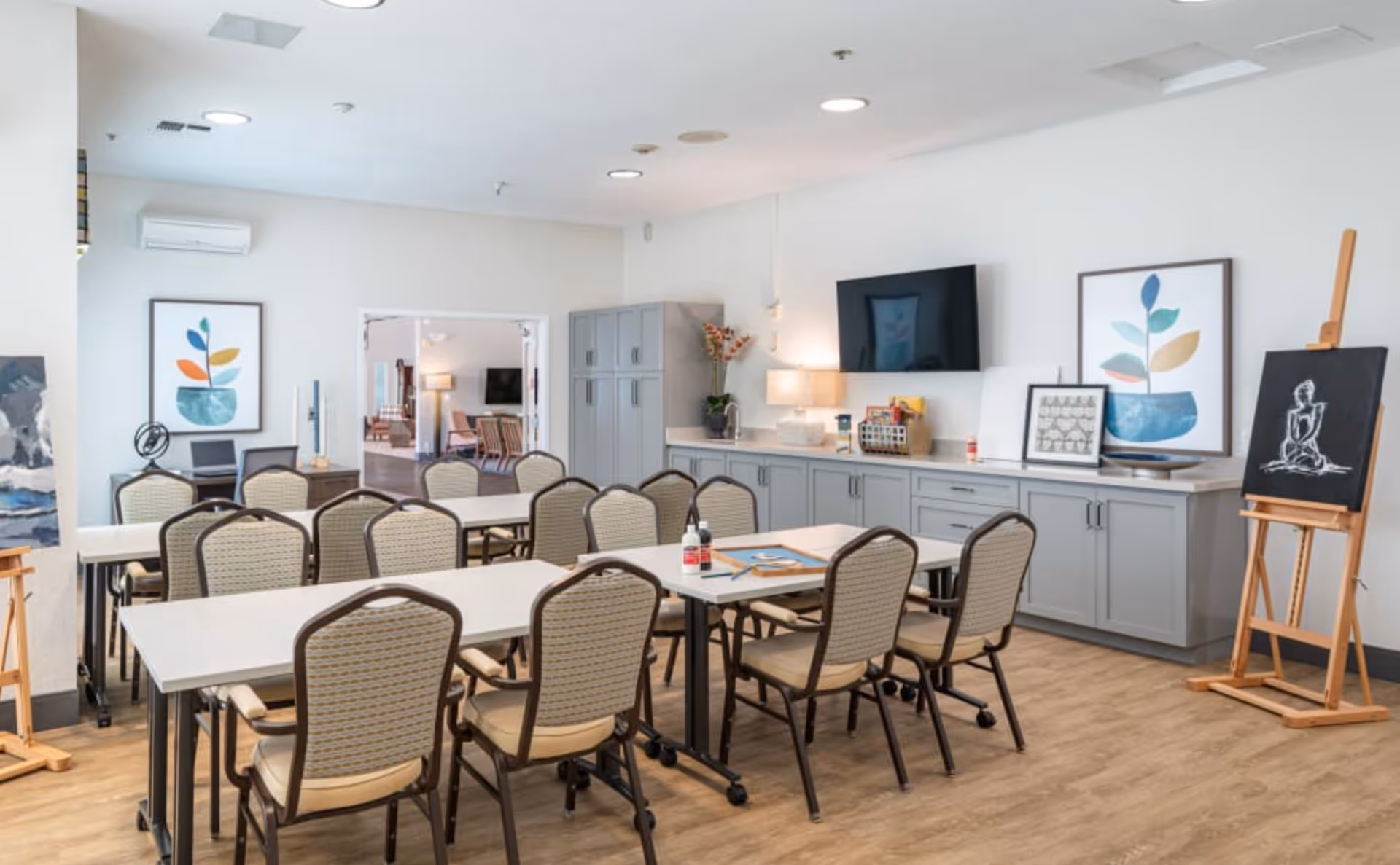 A bright, spacious room with several tables and chairs arranged for group activities or meetings. The room features light-colored walls, wood flooring, and modern cabinetry along one wall with a mounted flat-screen TV above. Decorative framed artwork and an easel with a drawing are also visible, creating a welcoming and creative atmosphere.