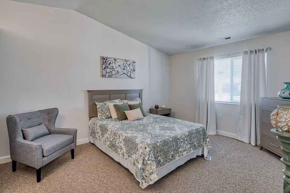 A neatly furnished bedroom with bed, nightstand, dresser, and an upholstered chair.
