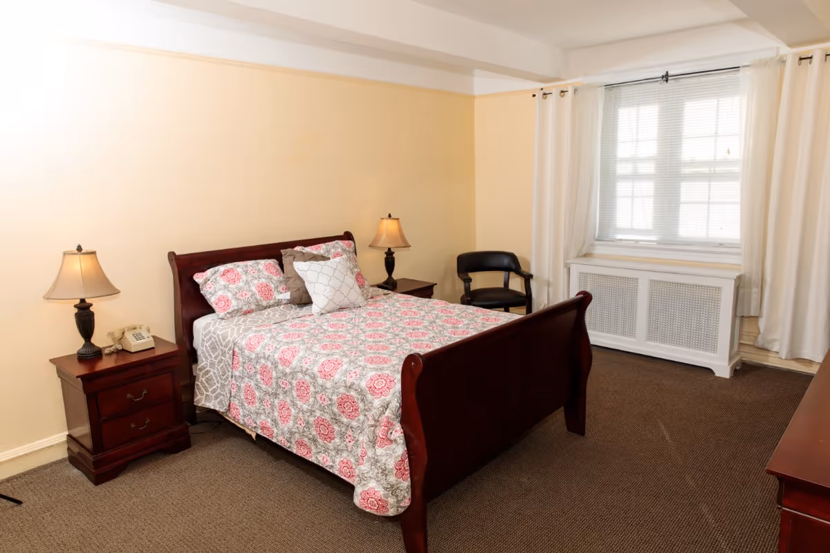 A bedroom with a wooden bed frame featuring a floral patterned bedspread and pillows. There are two bedside tables with lamps and a telephone on one side. A black chair is placed near a window with white curtains, and the room has beige walls and carpeted floor.