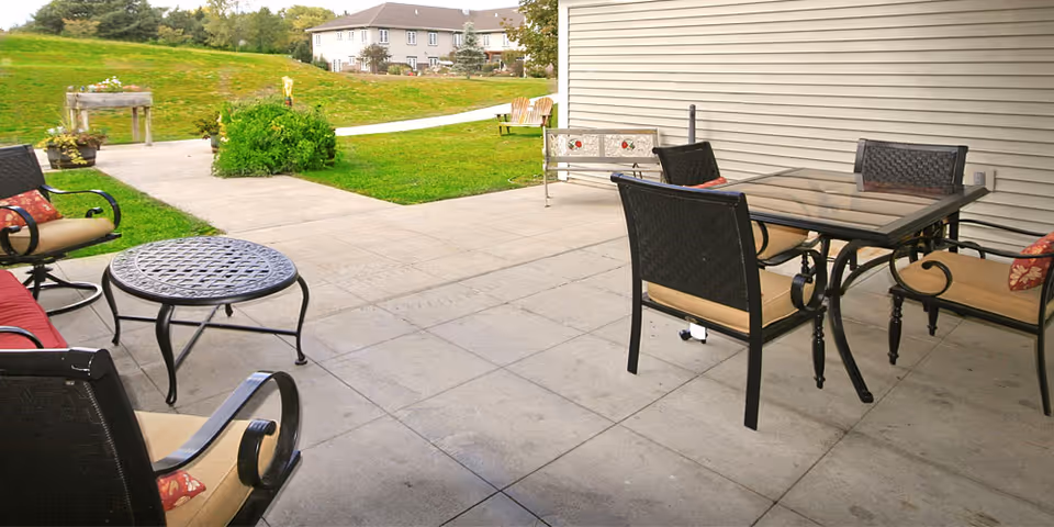 Outdoor patio with cushioned metal chairs around a glass-top dining table and a round metal coffee table, overlooking a grassy lawn and a distant building.