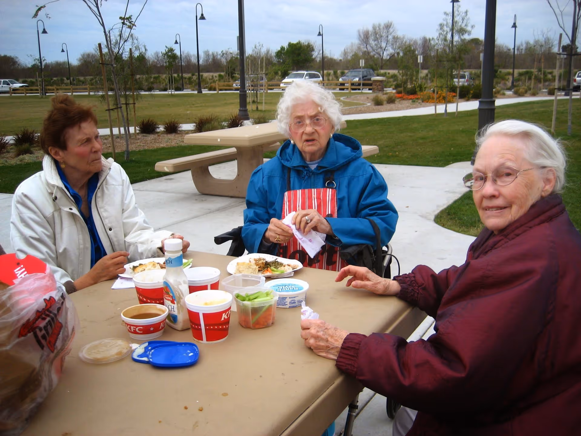 Three elderly women sitting around a picnic table outdoors, eating a meal with KFC containers and other food items on the table. They are dressed warmly with jackets and one woman is wearing an apron. The setting is a park-like area with grass, trees, and a paved walkway in the background.