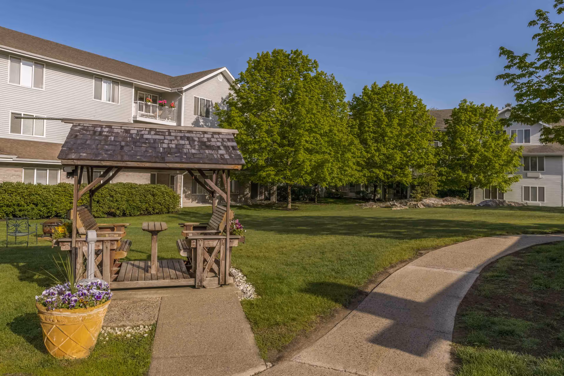 Wooden gazebo with seating on a grassy courtyard in front of a multi-story senior living building lined with trees.