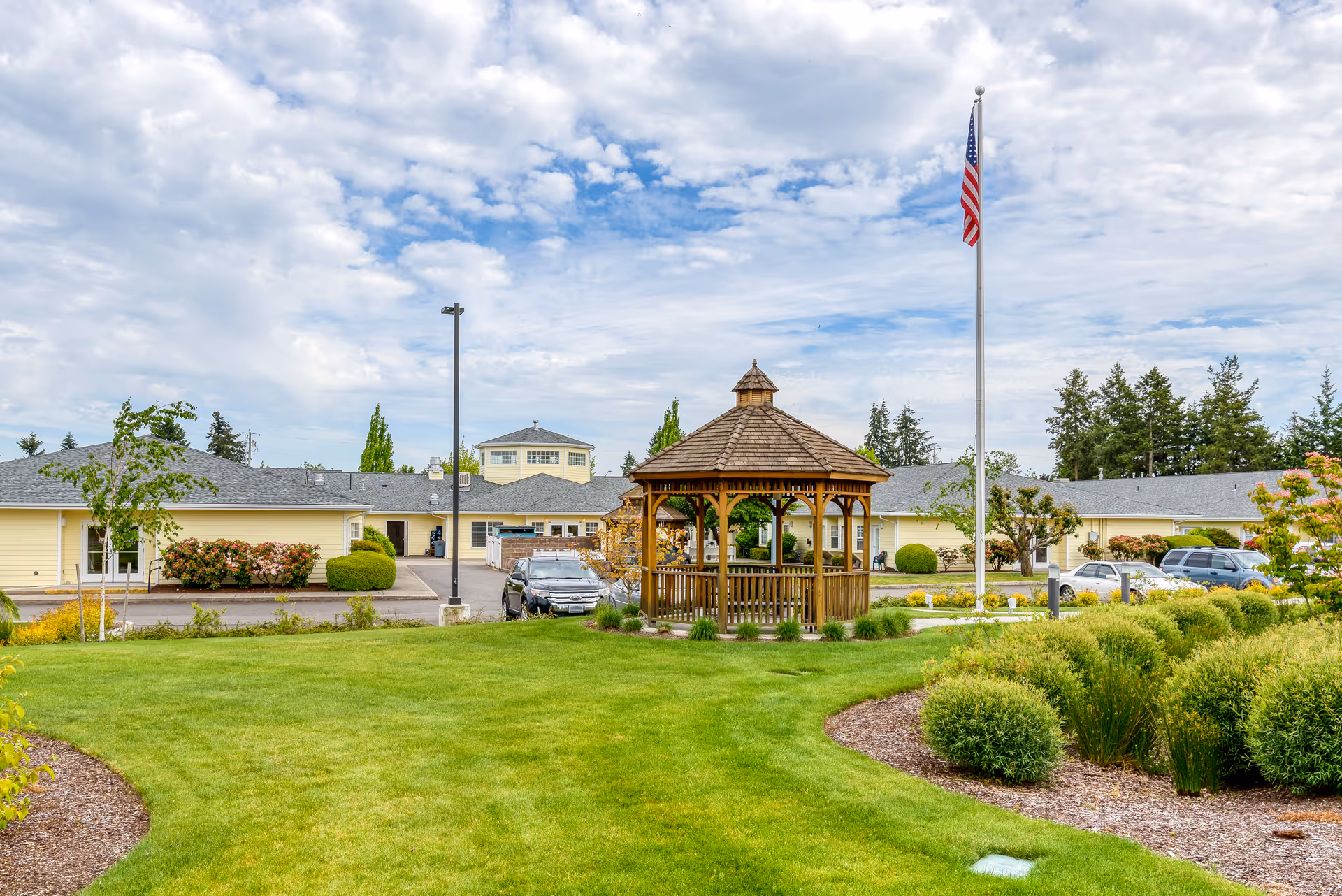 A well-maintained outdoor area of Easthaven Villa Assisted Living & Memory Care featuring a wooden gazebo in the center, surrounded by green grass, bushes, and landscaped garden beds. Several cars are parked near the single-story yellow building in the background, and an American flag flies on a flagpole to the right. The sky is partly cloudy.