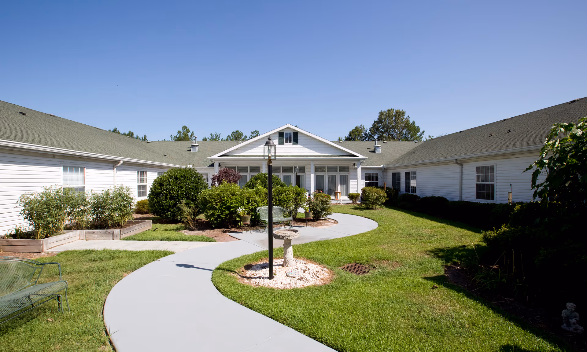A sunny outdoor courtyard area of a senior living facility with a curved concrete walkway, green grass, bushes, and a white building with green roof surrounding the courtyard. There is a black lamp post and a birdbath in the center of the walkway, along with a green metal bench on the left side.