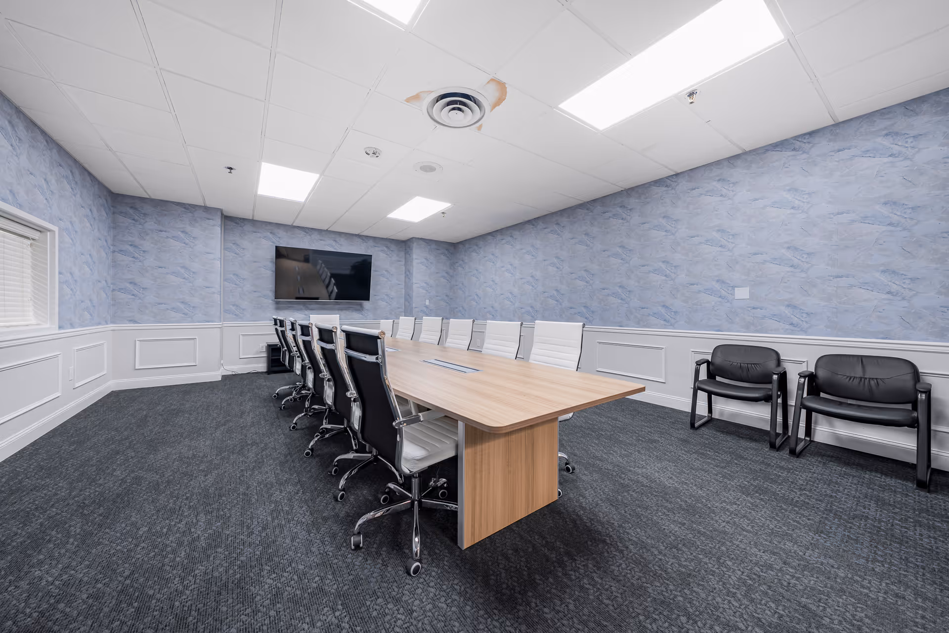 Conference room with a long wooden meeting table surrounded by office chairs and a wall-mounted TV.