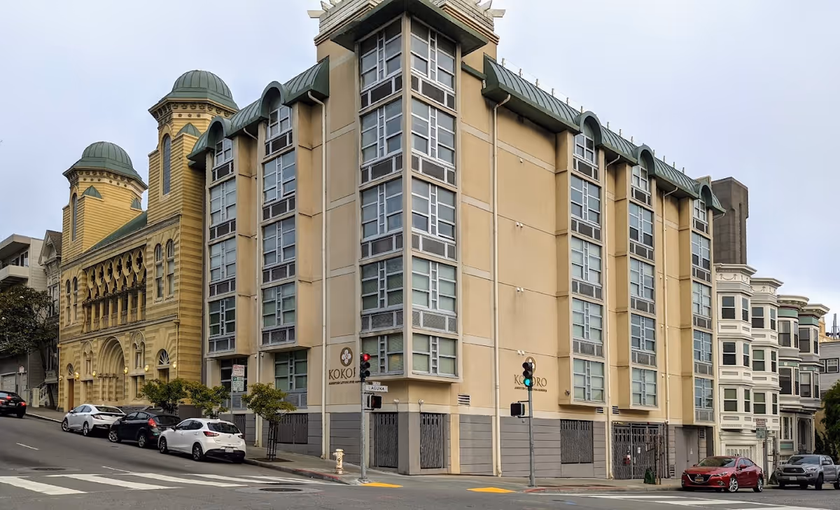 Exterior view of a multi-story building on a street corner with the name Kokoro Assisted Living Inc visible on the walls. The building has large windows and a beige facade with green roof accents. Several cars are parked along the street in front of the building, and there is a traffic light at the intersection.