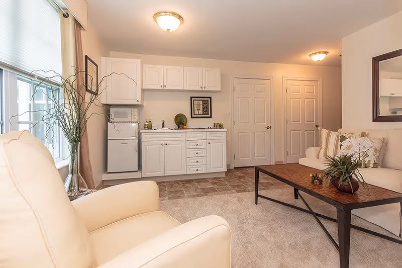 A cozy living room area with a beige armchair and a matching sofa adorned with decorative pillows. A wooden coffee table with a plant centerpiece sits in front of the sofa. The room features a small kitchenette with white cabinets, a mini fridge, microwave, and a sink. Two closed white doors are visible in the background, and a large window with beige curtains allows natural light to fill the space.