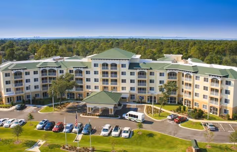 A large, multi-story senior living facility building with a green roof and beige exterior walls, surrounded by a parking lot with several cars and a white van. The building is set against a backdrop of dense green trees under a clear blue sky.