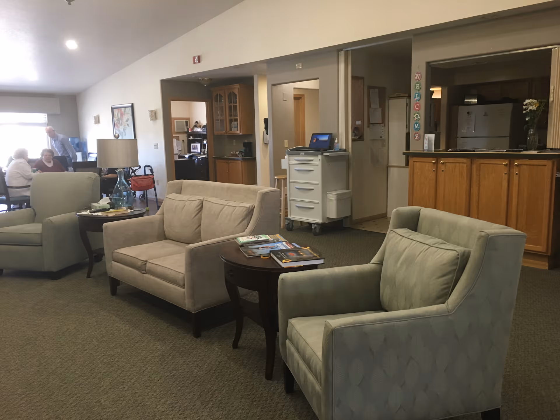 Communal lounge with armchairs, a loveseat, side tables and people sitting in the background near a kitchenette and reception area.