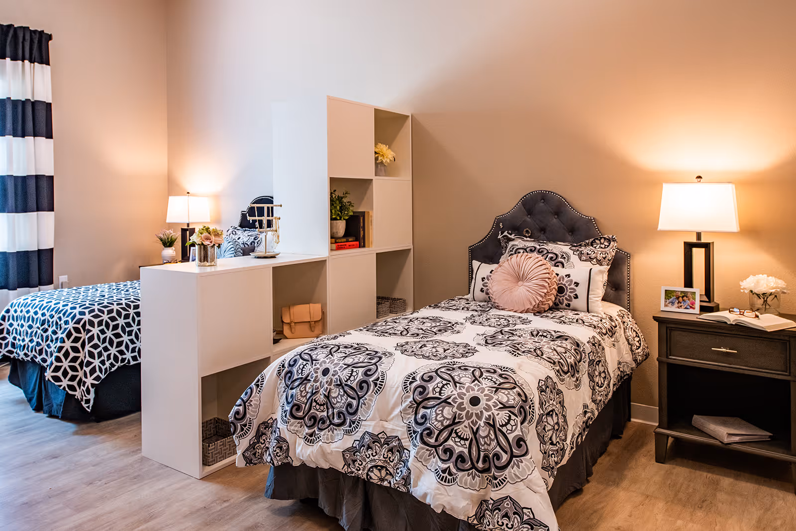 A cozy bedroom with two single beds separated by a white shelving unit. Each bed has patterned bedding and decorative pillows. There are two bedside tables with lamps, a small plant, a framed photo, and an open book. The room has wood flooring and a window with black and white striped curtains.