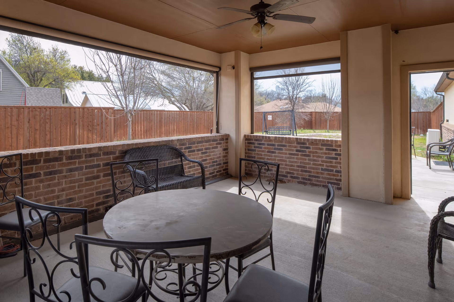 Covered outdoor patio area with a round metal table and four matching chairs, a black metal bench against a brick half-wall, and a view of a fenced yard with trees and neighboring houses in the background.