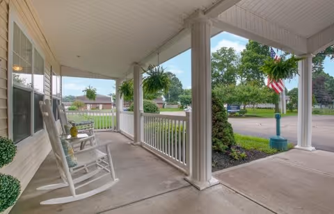 Covered porch area with white rocking chairs and hanging green plants, overlooking a landscaped garden and a street with trees and a flagpole displaying an American flag.