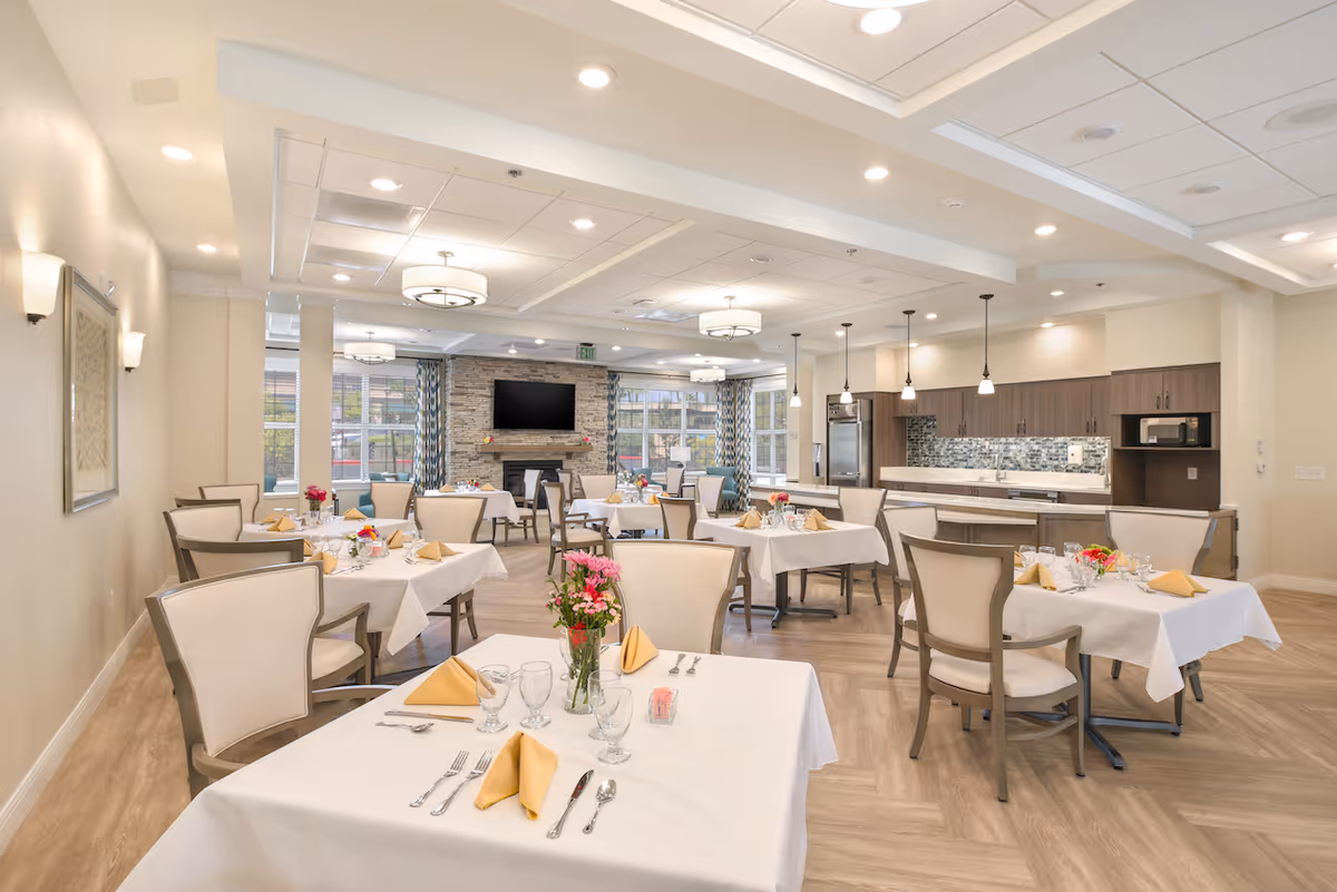 Bright, elegant dining room with multiple set tables and chairs, an open kitchen area, and a fireplace with TV in the background.
