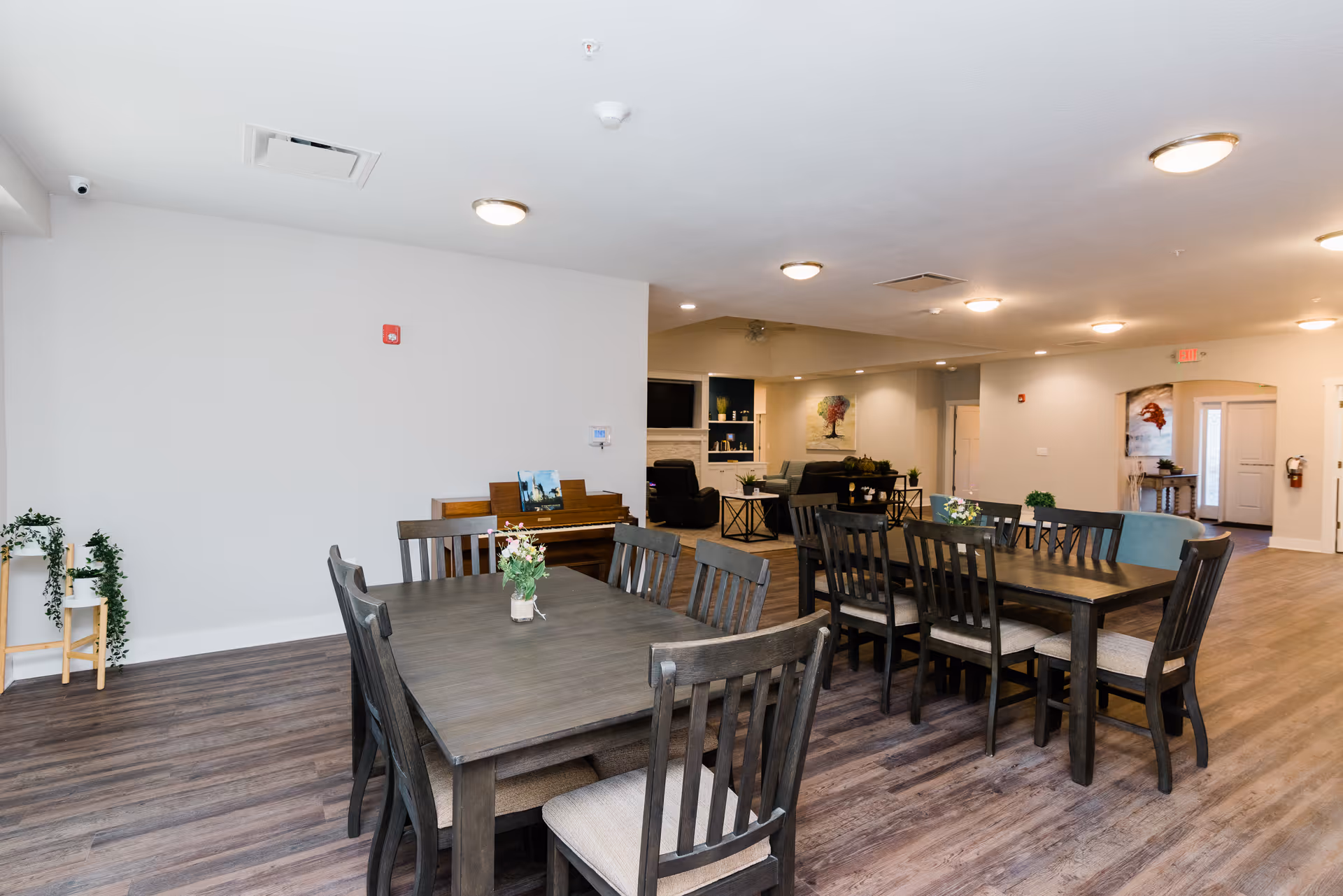 Open communal dining and lounge area with dark wood dining tables and chairs on wood-look floors and a seating area in the background.