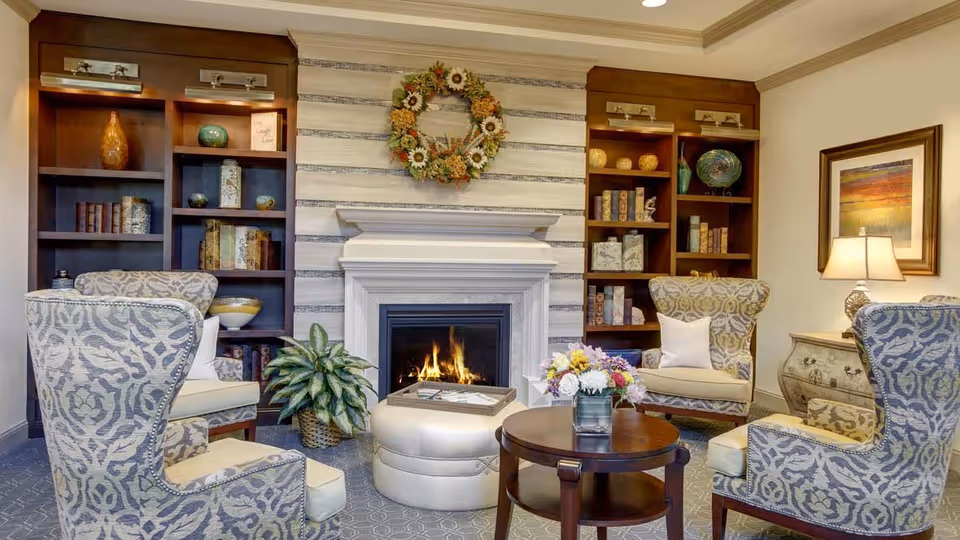 A cozy living room with four patterned armchairs arranged around a round wooden coffee table and a round ottoman. A fireplace with a floral wreath above it is centered between two wooden bookshelves filled with decorative items and books. A potted plant is placed near the fireplace, and a table lamp with a framed painting is visible on the right side.