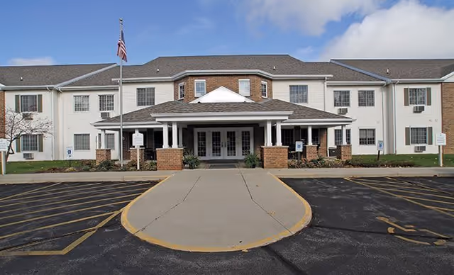 Front exterior view of a two-story senior living facility building with a covered entrance, an American flag on a flagpole, and a parking area in front under a partly cloudy sky.