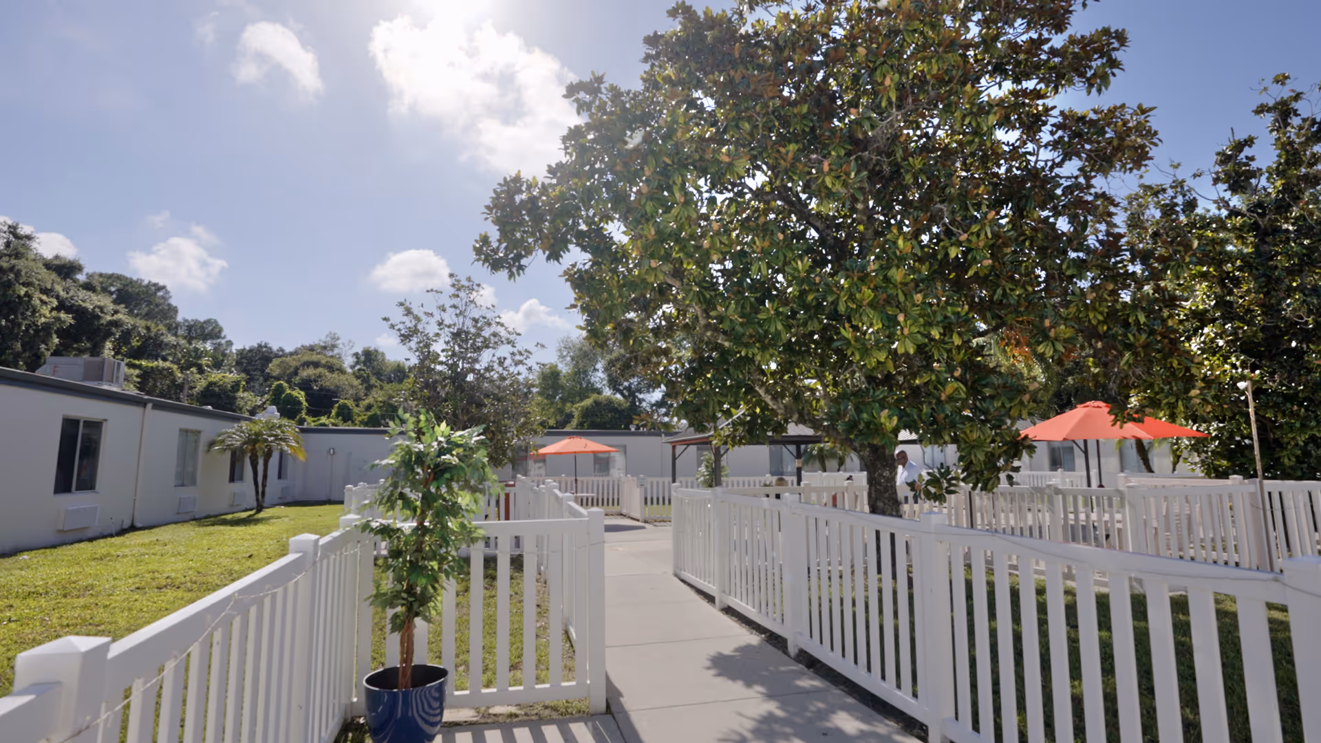 Outdoor courtyard area at Woodbridge Care Center with white fences, a paved walkway, green grass, trees, and red umbrellas providing shade over seating areas under a partly cloudy sky.