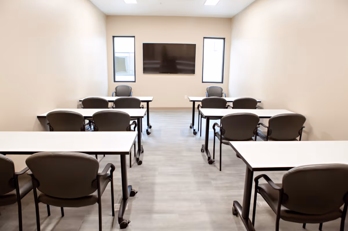 A small classroom or meeting room with several tables and chairs arranged in rows facing a wall-mounted flat screen TV. The room has light-colored walls, two narrow windows on either side of the TV, and a light gray floor.