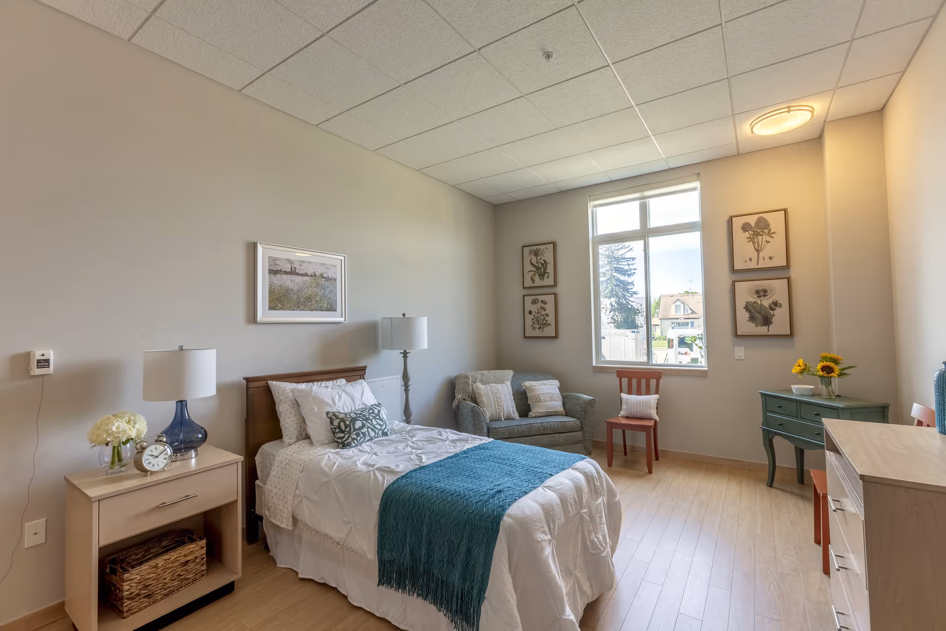 A bright and cozy bedroom in a senior living facility featuring a single bed with white bedding and a teal throw blanket, a wooden nightstand with a lamp, clock, and flowers, a small sofa with cushions, a wooden chair, a green dresser with sunflowers in a vase, and framed botanical prints on the walls. A large window lets in natural light, illuminating the light wood flooring and neutral-colored walls.