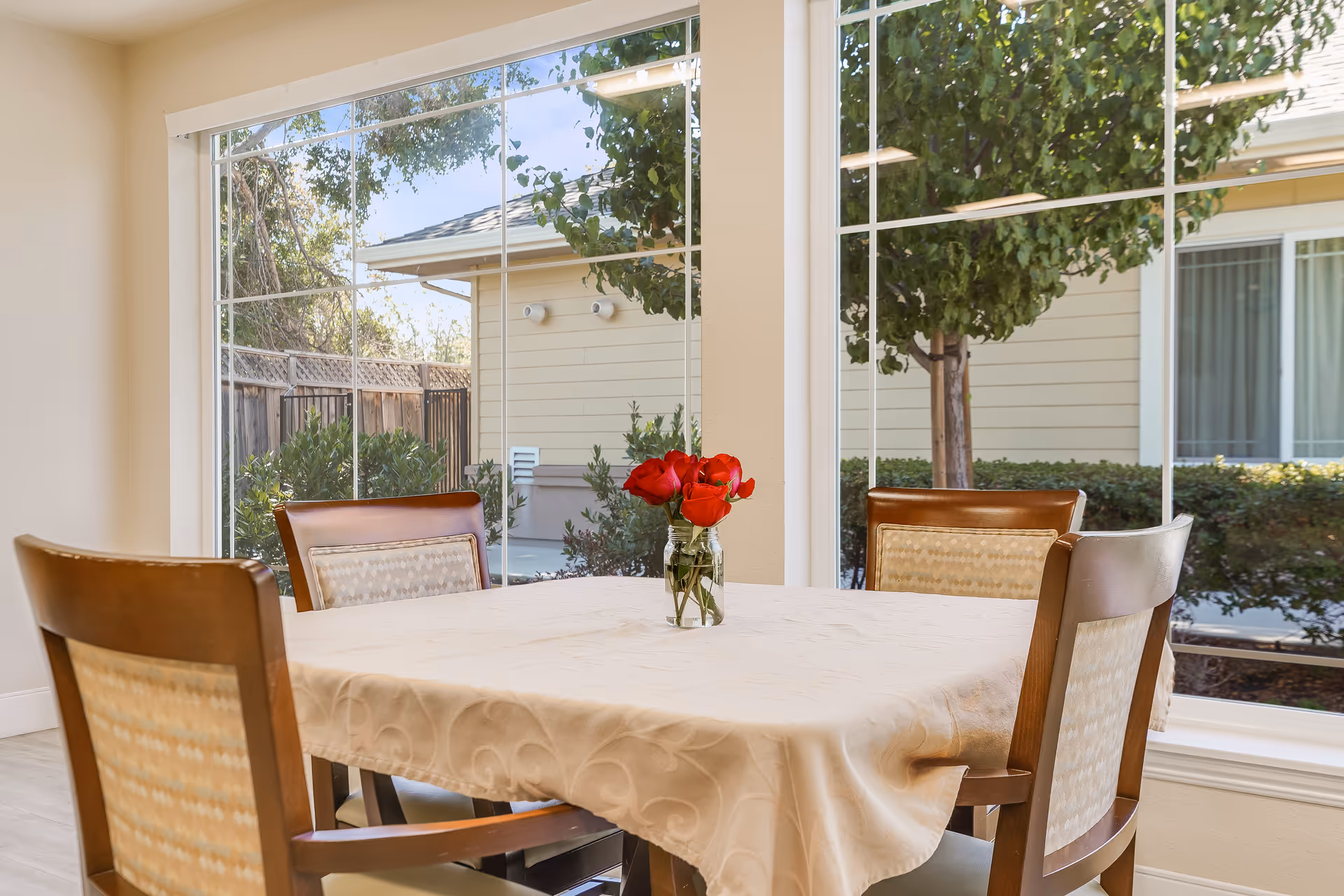 A dining table covered with a beige tablecloth and a small vase with red roses in the center, surrounded by four wooden chairs with patterned cushions. Large windows behind the table show an outdoor view with trees, bushes, and a building exterior.