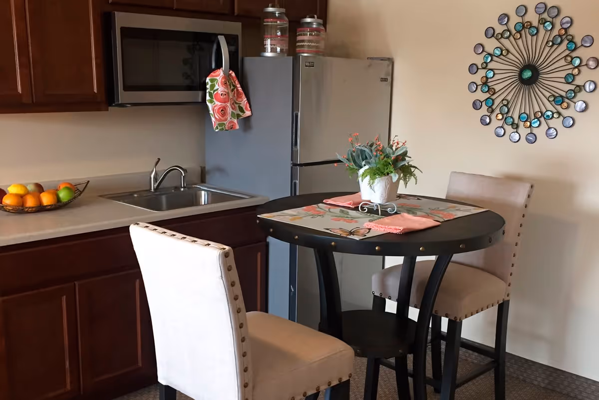 A small kitchen area with dark wooden cabinets, a stainless steel microwave, a sink, and a refrigerator. A round black table with two beige upholstered chairs is set with a floral placemat, pink napkins, and a small potted plant. A decorative wall piece with circular elements is mounted on the wall.