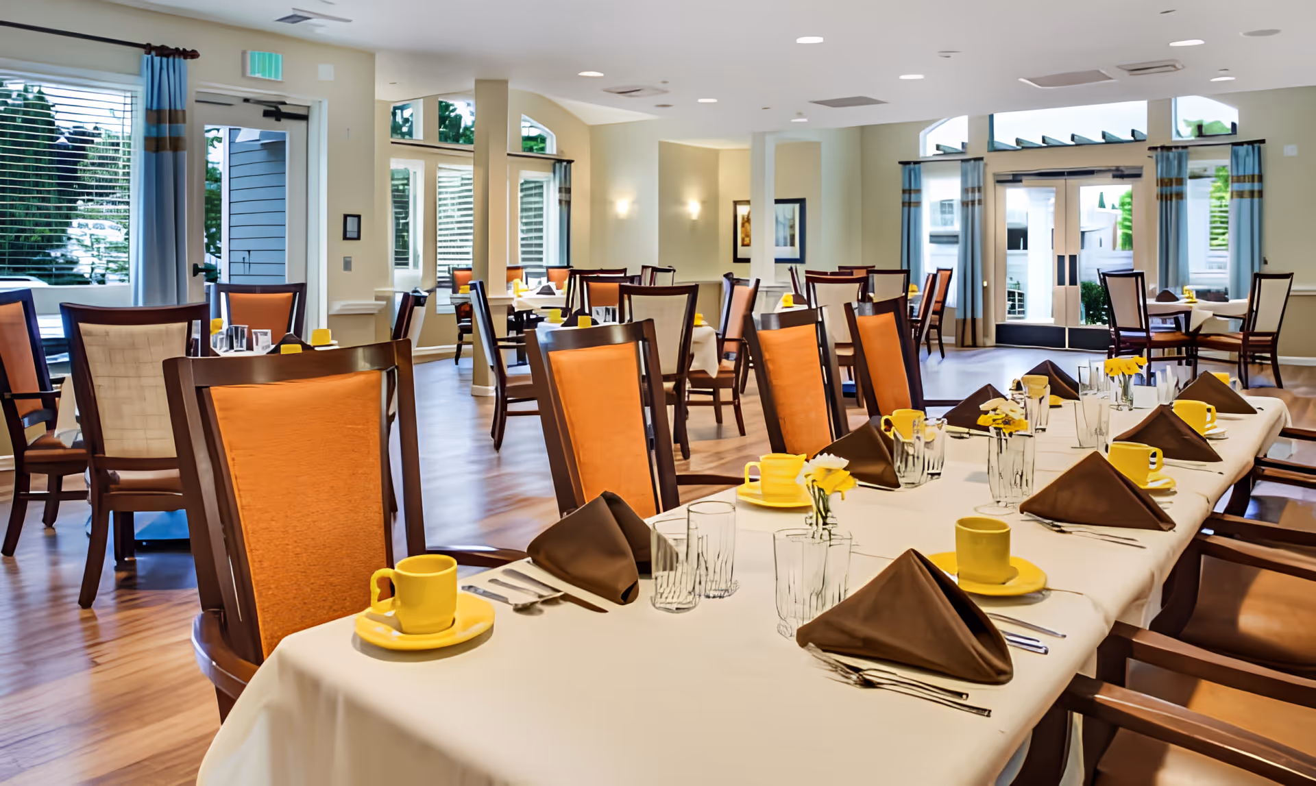 A bright and spacious dining room with multiple tables set with white tablecloths, brown folded napkins, yellow cups and saucers, clear drinking glasses, and small yellow flower centerpieces. The room has wooden chairs with orange and beige cushions, large windows with blue curtains, and a door leading outside.