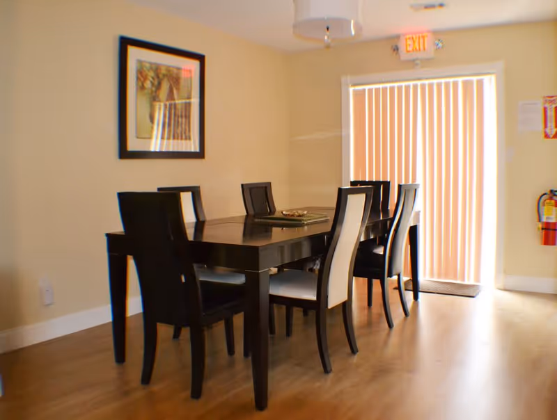 A dining area with a dark wooden table and six matching chairs with light-colored cushions. The room has light-colored walls, a framed picture on the wall, vertical blinds covering a glass door, and a fire extinguisher mounted on the wall near the door.
