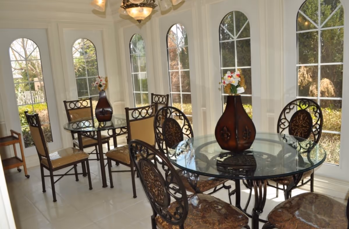 Sunlit dining area with glass-top tables, ornate metal chairs, and large arched windows.