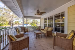 Covered outdoor patio area with wicker furniture including two armchairs, a loveseat, and a coffee table. The patio has a ceiling fan and overlooks a garden with trees and greenery.