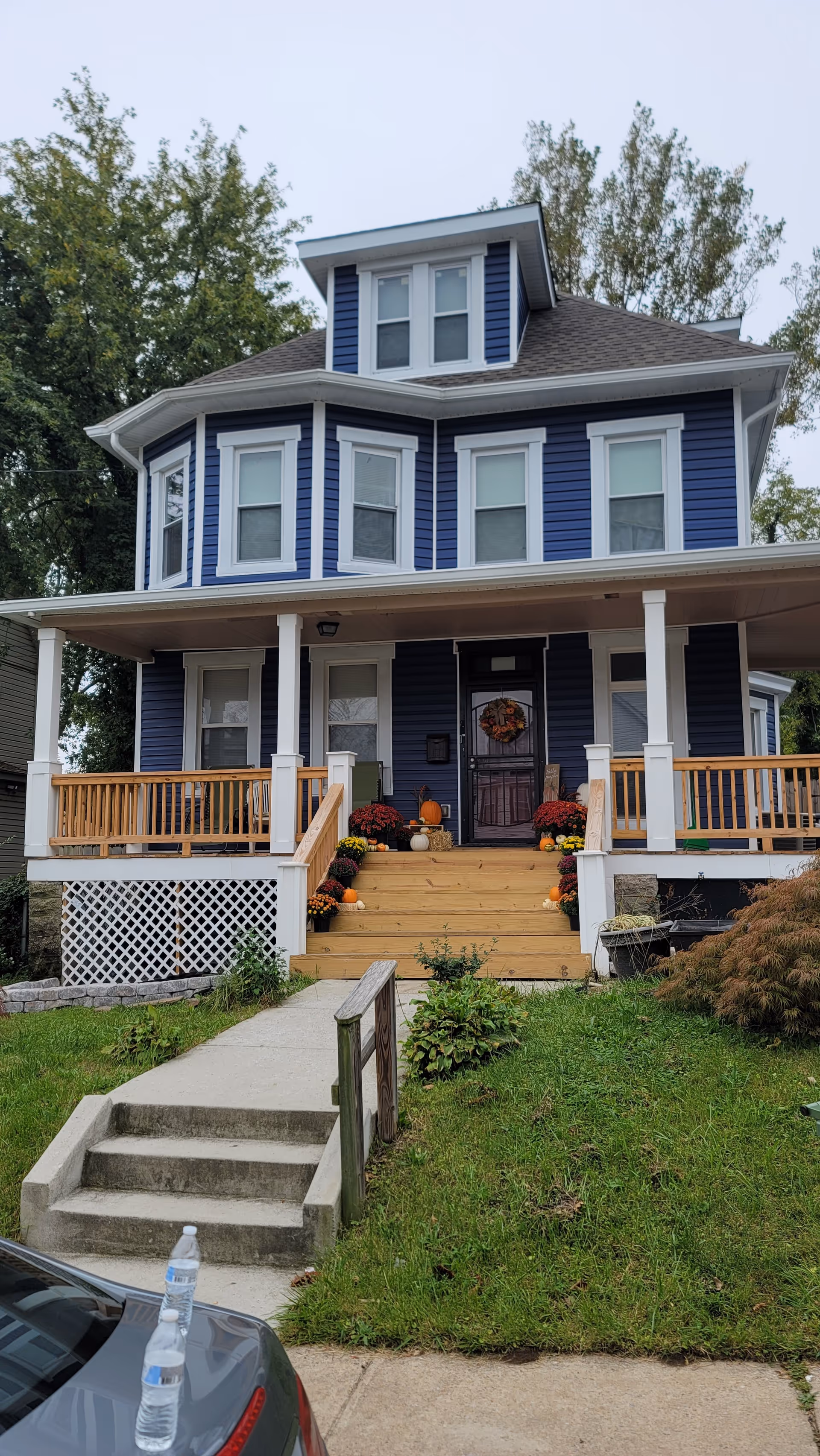 A two-story blue house with white trim and a front porch decorated with pumpkins and flowers. There are steps leading up to the porch from a concrete walkway, and a car with a water bottle on its trunk is partially visible in the foreground. Trees and grass surround the house.