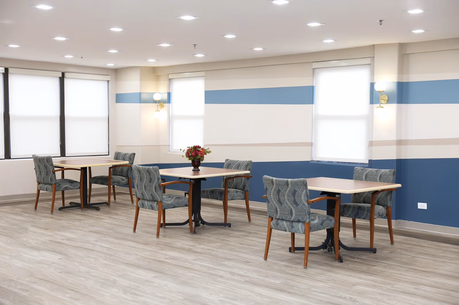 A bright and clean dining area with three square tables, each surrounded by four patterned chairs with wooden legs. The walls are painted with horizontal stripes in shades of blue and beige. There are three windows with white roller shades, and two wall-mounted light fixtures with round bulbs. The floor has light-colored wood-like flooring.