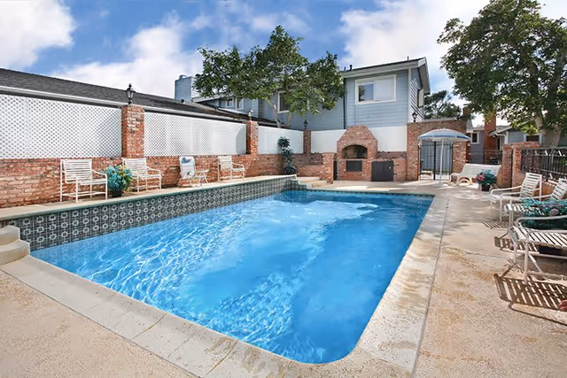 Outdoor swimming pool area with clear blue water surrounded by a concrete deck. There are several white chairs and small tables along the sides of the pool. A brick wall with lattice fencing and a built-in brick fireplace or grill is visible in the background, along with some trees and a two-story building under a partly cloudy sky.