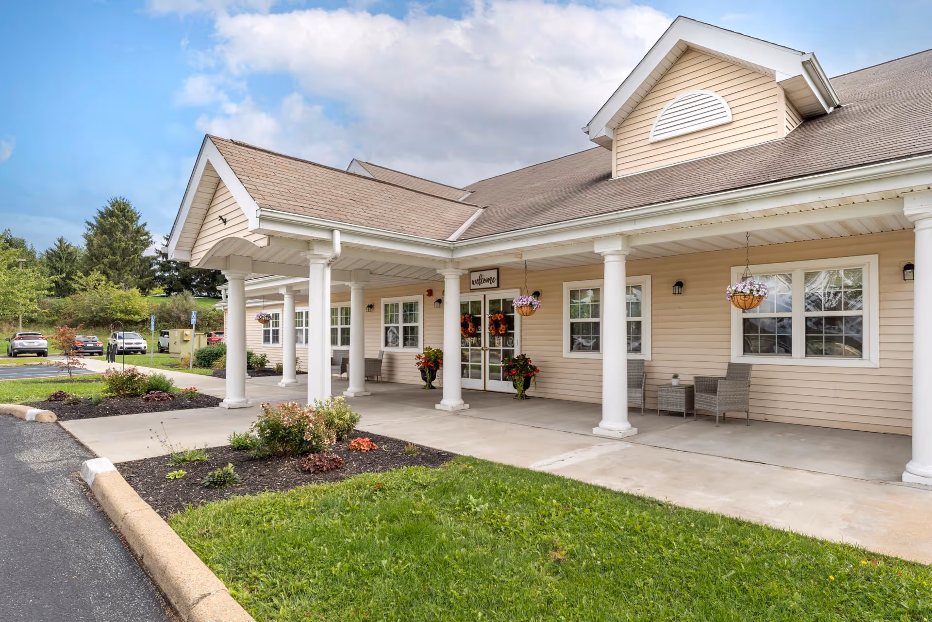 Covered front entrance of a beige senior living building with white columns, hanging flower baskets, seating, and a driveway.