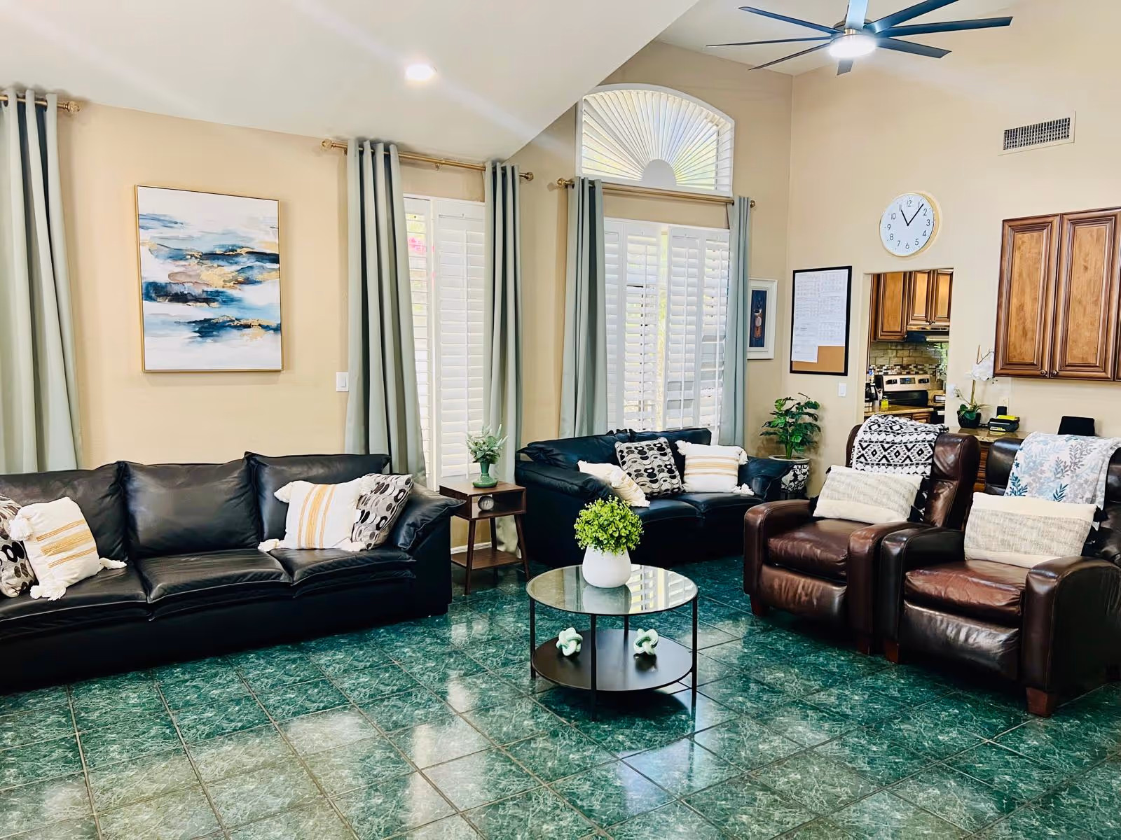 A bright and spacious living room with green tiled flooring, two black leather sofas, two brown leather armchairs with pillows, a round glass coffee table with a small green plant, large windows with white shutters and light gray curtains, a ceiling fan, and a kitchen area visible in the background with wooden cabinets and a wall clock.