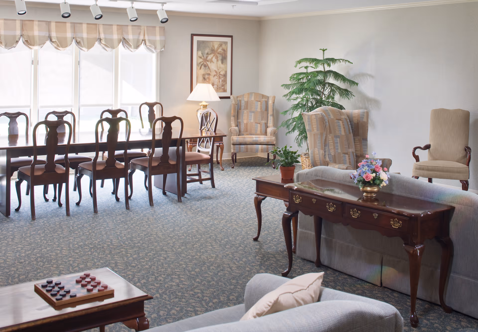 A well-lit common area in a senior living facility featuring a long wooden dining table with eight chairs near large windows with beige valances. The room also includes a seating area with upholstered armchairs, a sofa, a wooden console table with a flower arrangement, a side table with a lamp, a potted plant, and a checkers game on a small table in the foreground.