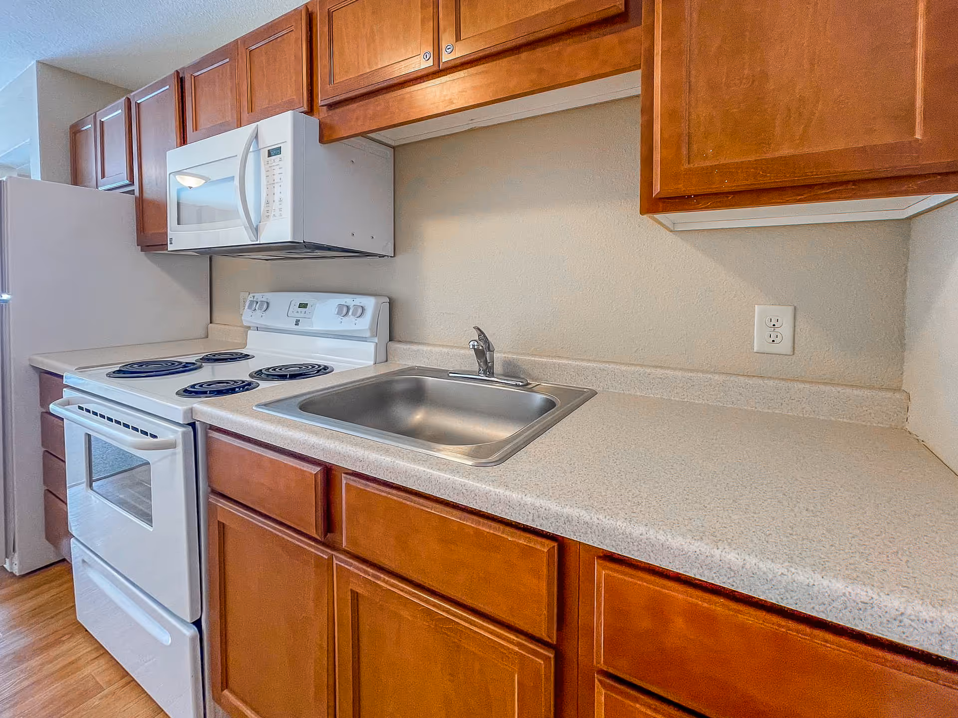 Interior view of a kitchen featuring wooden cabinets, a white electric stove with four coil burners, a white microwave mounted above the stove, a stainless steel sink with a single faucet, and a white refrigerator in the background.