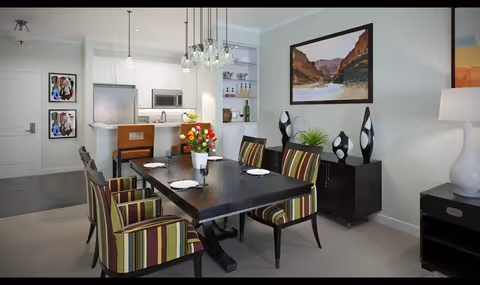 Open-plan dining area with a dark rectangular table set for four, striped upholstered chairs, decorative vases, and a view into a white kitchen.