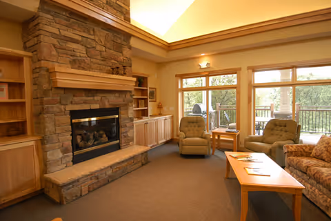 A cozy living room with a stone fireplace, wooden built-in shelves, two armchairs, a sofa, a wooden coffee table, and large windows overlooking a balcony with outdoor seating and greenery.